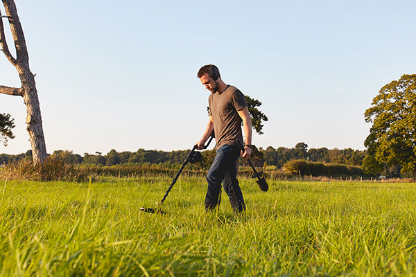 Man using a metal detector in a grassy field with trees in the background